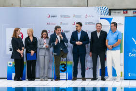 Pokaż zdjęcie: Zdjęcie przedstawia grupę siedmiu osób – czterech mężczyzn i trzech kobiet – stojących wzdłuż krawędzi basenu na ceremonii otwarcia lub wręczania nagród. Wszyscy są ubrani w stroje formalne, głównie ciemne garnitury i garsonki. Mężczyzna pośrodku, opierający się na jednej lasce i z widoczną protezą nogi, przemawia do mikrofonu. Po obu stronach stoją kolejni uczestnicy wydarzenia. W tle widoczne są loga sponsorów i instytucji, takich jak Ministerstwo Sportu i Turystyki oraz PFRON.