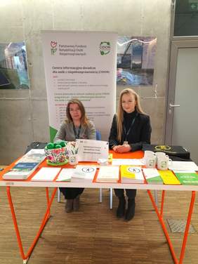 Pokaż zdjęcie: Przy stoliku na którym leżą ulotki, foldery, gadżety siedzą dwie kobiety z długimi włosami. W tle baner z logo CIDON