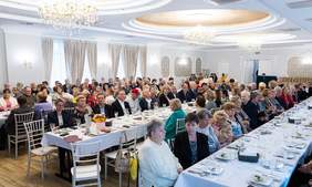 Pokaż zdjęcie: Zdjęcie przedstawia elegancką salę z licznymi uczestnikami siedzącymi przy długich, zastawionych stołach. W tle widoczne są dekoracje w stylu klasycznym z żyrandolami i zasłonami. Goście uczestniczą w wydarzeniu związanym z edukacją zdrowotną i promocją zdrowego stylu życia.