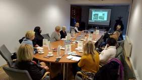 Pokaż zdjęcie: Zebrani goście siedzą przy stole. Na monitorze oglądają prezentację omawianą przez jednego z prelegentów.