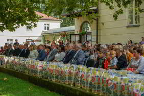 Ma trawie na niewysokim murku stoją ustawione w szeregu kolorowe reklamówki. Za nimi na tle jasnych budynków siedzi kilkudziesięciu uczestników wydarzenia