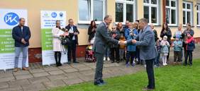 Na tle jasnego budynku stoi grupa ludzi, dorośli i dzieci, biją brawo. Przed nimi na pierwszym planie zdjęcia dwóch mężczyzn ściska sobie dłonie.