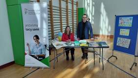 Pokaż zdjęcie: Przy stoisku informacyjnym PFRON znajdują sie dwie osoby: kobieta i mężczyzna. Na stoliku leżą różne informatory, poradniki, broszury, ulotki. W tle widać wyposażenie szkolnej sali gimnastycznej. 