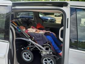 Pokaż zdjęcie: We wnętrzu auta znajduje się osoba siedząca na wózku inwalidzkim. Samochód jest w białym kolorze.  