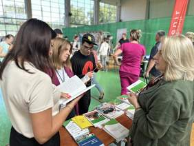 Pokaż zdjęcie: Przy stoliku na którym leżą informatory i ulotki stoją trzy osoby i pracownik Oddziału. W tle inne stanowiska.