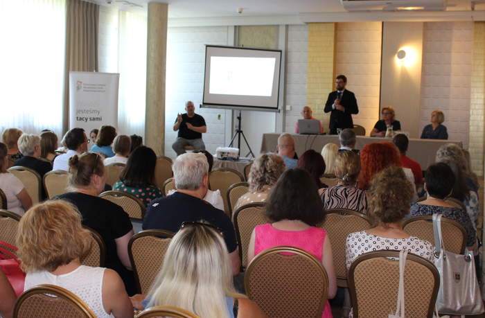 Pokaż zdjęcie: Sala szkoleniowa, na przodzie ekran z prezentacja, przy stole siedzi trzech prelegentów, słuchacze siedzą na krzesłach