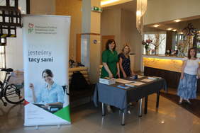 Pokaż zdjęcie: recepcja hotelu po lewej stronie widac stolik z ulotkami, za nim stoją dwie kobiety, po prawej stronie od stolika kobieta. Z lewej strony zdjęcia stoi rolap z logo PFRON