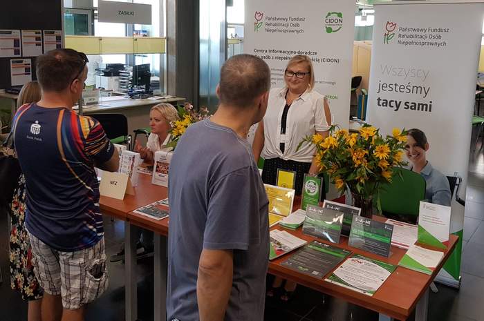 Pokaż zdjęcie: Jedna uśmiechnięta kobieta stoi na tle baneru PFRON oraz CIDON i udziela informacji mężczyźnie, który przyszedł do ZUS na Dzień Osób z Niepełnosprawnością. Na stoliku ułożone są materiały informacyjne promujące programy i zadania PFRON.
