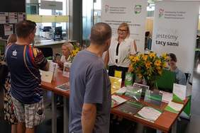 Pokaż zdjęcie: Jedna uśmiechnięta kobieta stoi na tle baneru PFRON oraz CIDON i udziela informacji mężczyźnie, który przyszedł do ZUS na Dzień Osób z Niepełnosprawnością. Na stoliku ułożone są materiały informacyjne promujące programy i zadania PFRON.