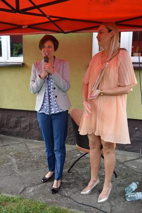 Pokaż zdjęcie: Dwie kobiety stoją pod namiotem. Jedna z nich przemawia do mikrofonu, natomiast druga trzyma w dłoniach rękodzieło oprawione w ramkę. W tle widoczny jest fragment budynku.