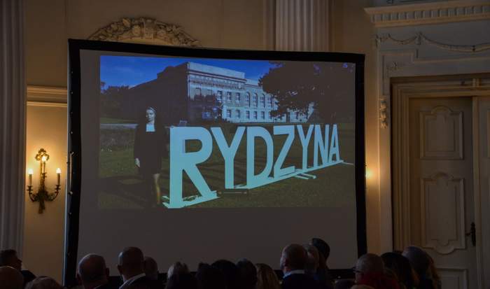 Pokaż zdjęcie: Na dużym ekranie wewnątrz sali zamkowej w Rydzynie wyświetlany jest film, na którym widoczny jest niniejszy zamek z zewnątrz oraz kobieta stojąca przy dużym napisie „Rydzyna” ustawionym na trawniku. Film oglądają osoby znajdujące się w sali.