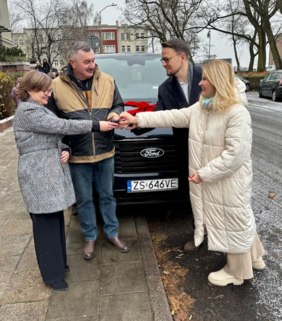 Pokaż zdjęcie: Cztery osoby stoją na chodniku obok czarnego vana marki Ford (z widoczną tablicą rejestracyjną ZS 646VE) z czerwoną kokardą na masce. Dwie kobiety i dwóch mężczyzn uczestniczą w ceremonii przekazania kluczyków. Kobieta w długiej, beżowej pikowanej kurtce i mężczyzna w dwukolorowej kurtce roboczej trzymają kluczyki, symbolicznie je sobie przekazując. Obok nich stoi drugi mężczyzna w ciemnym płaszczu i kobieta w szarym płaszczu. Tło stanowią budynki mieszkalne i ulica.