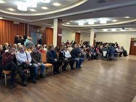 Pokaż zdjęcie: W sali są ustawione w rzędach krzesła. Siedzą na nich ludzie. Niektórzy siedzą na wózkach inwalidzkich. W tle widać stoły z poczęstunkiem. 