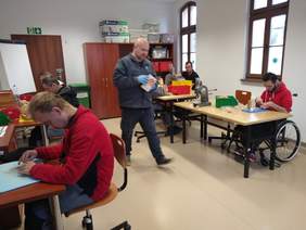 Pokaż zdjęcie: 6 osób w skupieniu pracuje nad przygotowaniem naturalnej podpałki: złożeniem drewnianych patyczków, przycinaniem elementów gilotyną. Jeden z pracujących młodych mężczyzn jest na wózku.