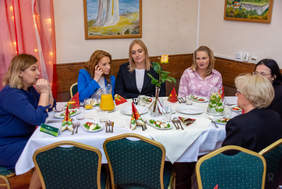 Pokaż zdjęcie: Grupa kobiet siedzi przy stole świątecznym, na środku którego stoi wazon z żółtą różą. Każda z uczestniczek ma przed sobą nakrycie z sałatką i napojami. W tle widoczne dekoracje i obrazy.