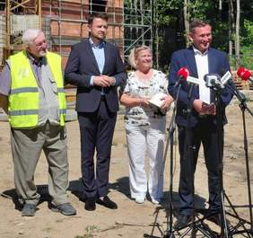 Fotografia plenerowa przedstawiająca cztery osoby stojące w rzędzie podczas konferencji prasowej, z fragmentem piątej osoby (po prawej) trzymającej mikrofon. Od lewej: starszy mężczyzna w odblaskowej kamizelce z kaskiem pod pachą, młodszy mężczyzna w garniturze, kobieta w białych spodniach z kaskiem w dłoniach, i drugi mężczyzna w garniturze przemawiający do mikrofonów. W tle widać fragmenty rusztowań i piaszczystego terenu budowy pod błękitnym niebem.