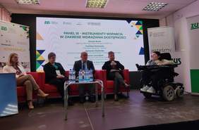 Pokaż zdjęcie: Pięciu panelistów bierze udział w dyskusji na konferencji ZUS. Cztery osoby siedzą na czerwonych fotelach, a jedna uczestniczka na wózku elektrycznym. Mężczyzna trzyma mikrofon i mówi, pozostali słuchają. W tle duży ekran z tytułem panelu i nazwiskami prelegentów, banery ZUS i PFRON. Na stoliku stoją butelki wody. Po lewej stronie widoczny jest ekran z tłumaczem języka migowego.