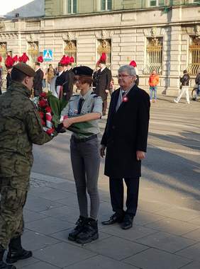 Pokaż zdjęcie: ulica Piotrkowska, na której stoją harcerze oraz Dyrektor Oddziału Łódzkiego PFRON ubrany jest w granatowy płaszcz przypięty ma kotylion w tle  defilada kampanii honorowych 