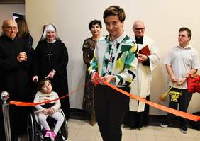 Pokaż zdjęcie: Kobieta blondynka z krótkimi włosami stoi przodem do zdjęcia w ręku trzyma szarfę i nożyczki. W tle siostry zakonne oraz wychowankowie ośrodka