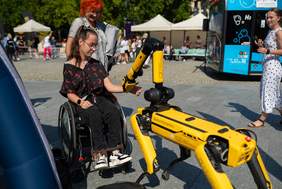 Pokaż zdjęcie: Robot przypominający psa podszedł do dziewczynki na wózku inwalidzkim, robot jest w kolorze żółtym.