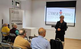 Pokaż zdjęcie: Uczestnicy konferencji siedzą na krzesłach i patrzą na przemawiającą na środku sali kobietę