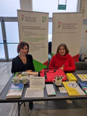 Pokaż zdjęcie: Przy stoliku na którym leżą ulotki siedzą dwie kobiety, w tle stoją rollupy z logo PFRON i CIDON