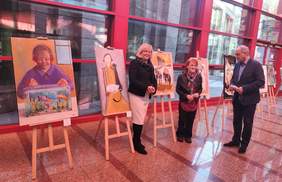 Pokaż zdjęcie: Dwie kobiety i jeden mężczyzna stoją uśmiechnięci, rozmawiają. W tle obrazy i grafiki. Prace ustawione są na drewnianych sztalugach.