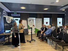 Pokaż zdjęcie: Dwie kobiety wygłaszają prelekcję w sali konferencyjnej, na krzesłach siedzą zaproszeni goście.