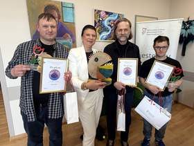 Do zdjęcia pozuje kobieta i trzech mężczyzn. Panowie - laureaci trzymają dyplomy i nagrody. Kobieta trzyma drewnianą kolorową rzeźbę która została nagrodzona.