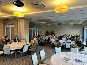 Pokaż zdjęcie: Sala konferencyjna. Pod sufitem wiszą okrągłe lampy. W Sali pięć dużych okrągłych stołów przy których siedzą ludzie. Na Stołach materiały prasowe, kartki długopisy i filiżanki. W sali są jasne ściany wykonane z drewna. Dywan w kratę na 