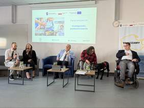 Pokaż zdjęcie: Pięcioro uczestników siedzi na scenie na fotelach, przed nimi małe stoliki z wodą i mikrofonami. Dwie osoby poruszają się na wózkach. W tle wyświetlona prezentacja „Panel ekspercki – Dostępność, podsumowanie”.