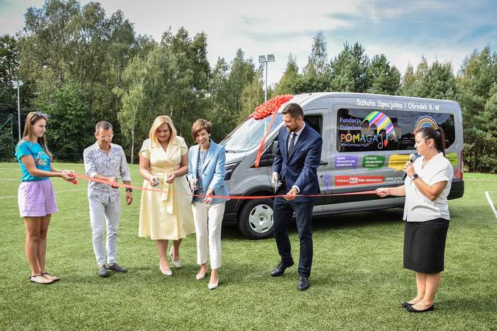 Pokaż zdjęcie: Dwie kobiety stoją na boisku i trzymają rozciągniętą wstęgę, do której przecięcia przymierzają się cztery inne osoby. Z tyłu znajduje się bus obklejony naklejkami z różnymi informacjami, a za nim rząd drzew liściastych i iglastych.