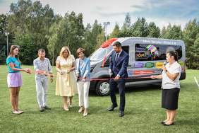 Pokaż zdjęcie: Dwie kobiety stoją na boisku i trzymają rozciągniętą wstęgę, do której przecięcia przymierzają się cztery inne osoby. Z tyłu znajduje się bus obklejony naklejkami z różnymi informacjami, a za nim rząd drzew liściastych i iglastych.