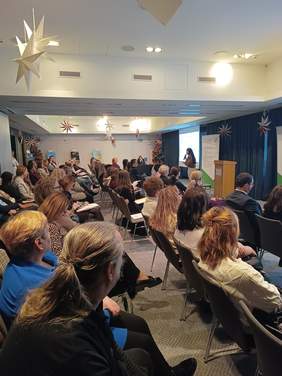 Pokaż zdjęcie: Zdjęcie sali konferencyjnej, na której kilkadziesiąt osób siedzi na krzesłach. Uczestnicy konferencji skierowani są w stronę mównicy przy, której stoi kobieta i przemawia z mikrofonem w ręku. Na ścianie za nią znajduje się ekran z wyświetlonym slajdem prezentacji. W pomieszczeniu wiszą ozdobne gwiazdy z papieru. 