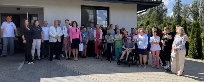 Pokaż zdjęcie: Liczna grupa uczestników, pracowników i zaproszonych gości pozuje do wspólnego zdjęcia przed budynkiem placówki. Wśród zgromadzonych są osoby z niepełnosprawnościami, w tym osoby poruszające się na wózkach i o kulach.