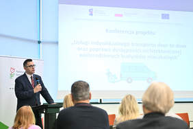 Pokaż zdjęcie: mężczyzna w ciemnej marynarce, okularach przemawia do mikrofonu