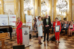 Pokaż zdjęcie: Dwie kobiety i mężczyzna stoją na tle zgromadzonych gości. Jedna z nich trzyma w ręku statuetkę i przygotowuje sie do jej wręczenia laureatowi