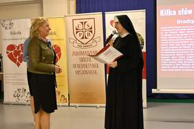 Pokaż zdjęcie: Na scenie stoi kobieta i zakonnica. Kobieta blondynka w zielonej ramonesce odbiera dyplom od siostry zakonnej.