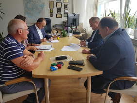 Przy stole konferencyjnym, z lewej strony dwóch mężczyzn i między nimi mało widoczna kobieta, z prawej dwóch mężczyzn, na stole dokumenty, pieczątki