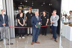 Pokaż zdjęcie: Wewnątrz kawiarni na środku stoi mężczyzna w garniturze trzyma zieloną teczkę przemawia za nim stoją pracownicy kawiarni 