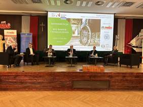 Pokaż zdjęcie: Na podwyższeniu stoją fotele ułożone w półokrąg. Na nich siedzą elegancko ubrani starsi ludzie. Za nimi telebim z napisem Konferencja bez barier i wykluczenia obok napisu zdjęcie wózka inwalidzkiego.