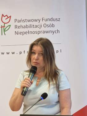 Pokaż zdjęcie: Kobieta z długimi włosami trzyma w ręku mikrofon, za nią stoją rollupy z logo PFRON.