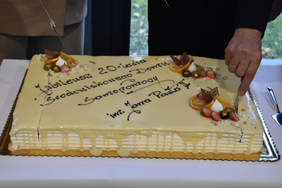 Pokaż zdjęcie: Na stoliku przykrytym białym obrusem znajduje się udekorowany owocami i bitą śmietaną tort wraz z napisem: „Jubileusz 20-lecia Środowiskowego Domu Samopomocy im. Jana Pawła II w Liskowie”. Tort został już częściowo przekrojony z obu stron.