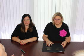 Pokaż zdjęcie: Przy stole konferencyjnym siedzą dwie kobiety w okularach, ubrane w ciemne stroje, na stole leżą umowy. 