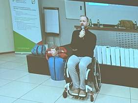 Mężczyzna w czarnym golfie i jasnych spodniach, siedzi na wózku, w ręce trzyma mikrofon