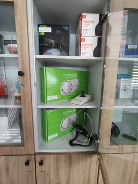 Pokaż zdjęcie: Przeszklona szafka z różnymi urządzeniami technologicznymi. Na górnej półce są słuchawki Shure i modele z funkcją redukcji hałasu. Na środkowej i dolnej półce znajdują się zielone pudełka z klawiaturami orbiTouch.