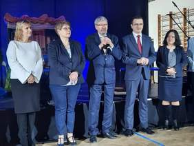 Pokaż zdjęcie: Na scenie stoi pięć osób: dwie kobiety i trzech mężczyzn. Pan stojący w środku trzyma w dłoniach mikrofon, do którego przemawia. Wszyscy są elegancko ubrani. 