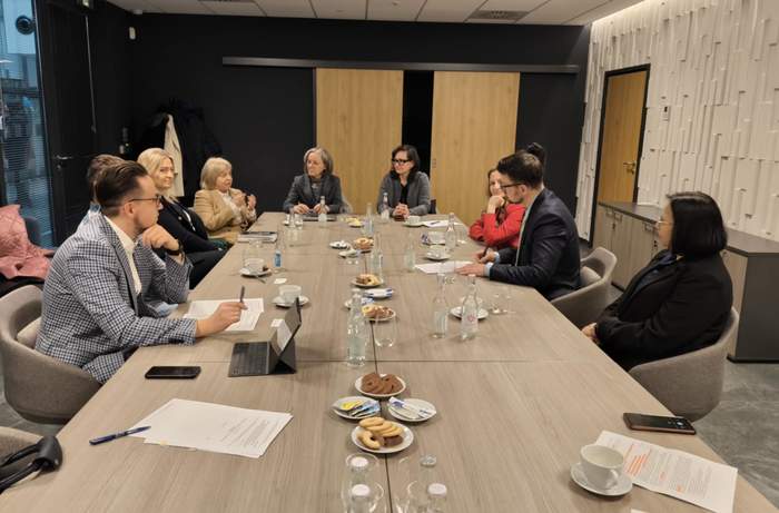 Pokaż zdjęcie: Grupa ośmiu osób siedzi przy długim stole konferencyjnym w nowoczesnym biurze. Uczestnicy są elegancko ubrani i zaangażowani w rozmowę, a na stole znajdują się dokumenty, filiżanki, butelki wody oraz talerze z ciastkami. W tle widać drewniane drzwi, ciemne ściany oraz białą, dekoracyjną panelową ścianę. Atmosfera sugeruje formalne spotkanie biznesowe lub naradę.