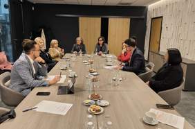 Pokaż zdjęcie: Grupa ośmiu osób siedzi przy długim stole konferencyjnym w nowoczesnym biurze. Uczestnicy są elegancko ubrani i zaangażowani w rozmowę, a na stole znajdują się dokumenty, filiżanki, butelki wody oraz talerze z ciastkami. W tle widać drewniane drzwi, ciemne ściany oraz białą, dekoracyjną panelową ścianę. Atmosfera sugeruje formalne spotkanie biznesowe lub naradę.