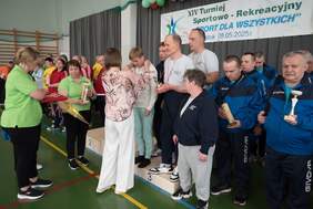 Pokaż zdjęcie: Grupa osób otrzymuje medale i puchary podczas ceremonii wręczenia nagród na turnieju sportowym. Na podium stoją uczestnicy w sportowych strojach, a osoby w zielonych koszulkach wręczają trofea. W tle widoczny jest baner z nazwą wydarzenia oraz datą: 8.05.2025 r.